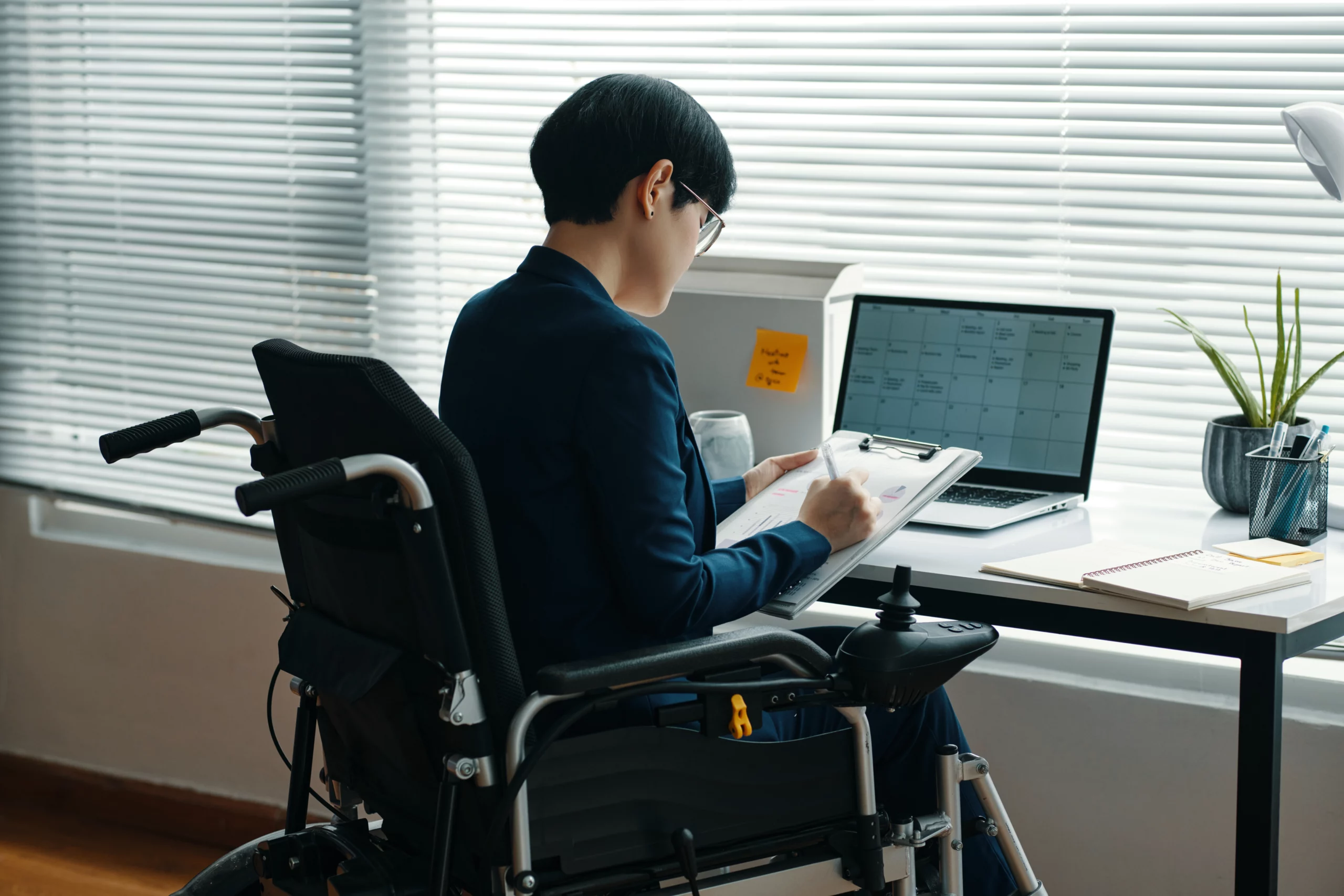 Professional in a wheelchair wearing glasses and a suit, writing on a clipboard at a desk with a laptop and notebooks.