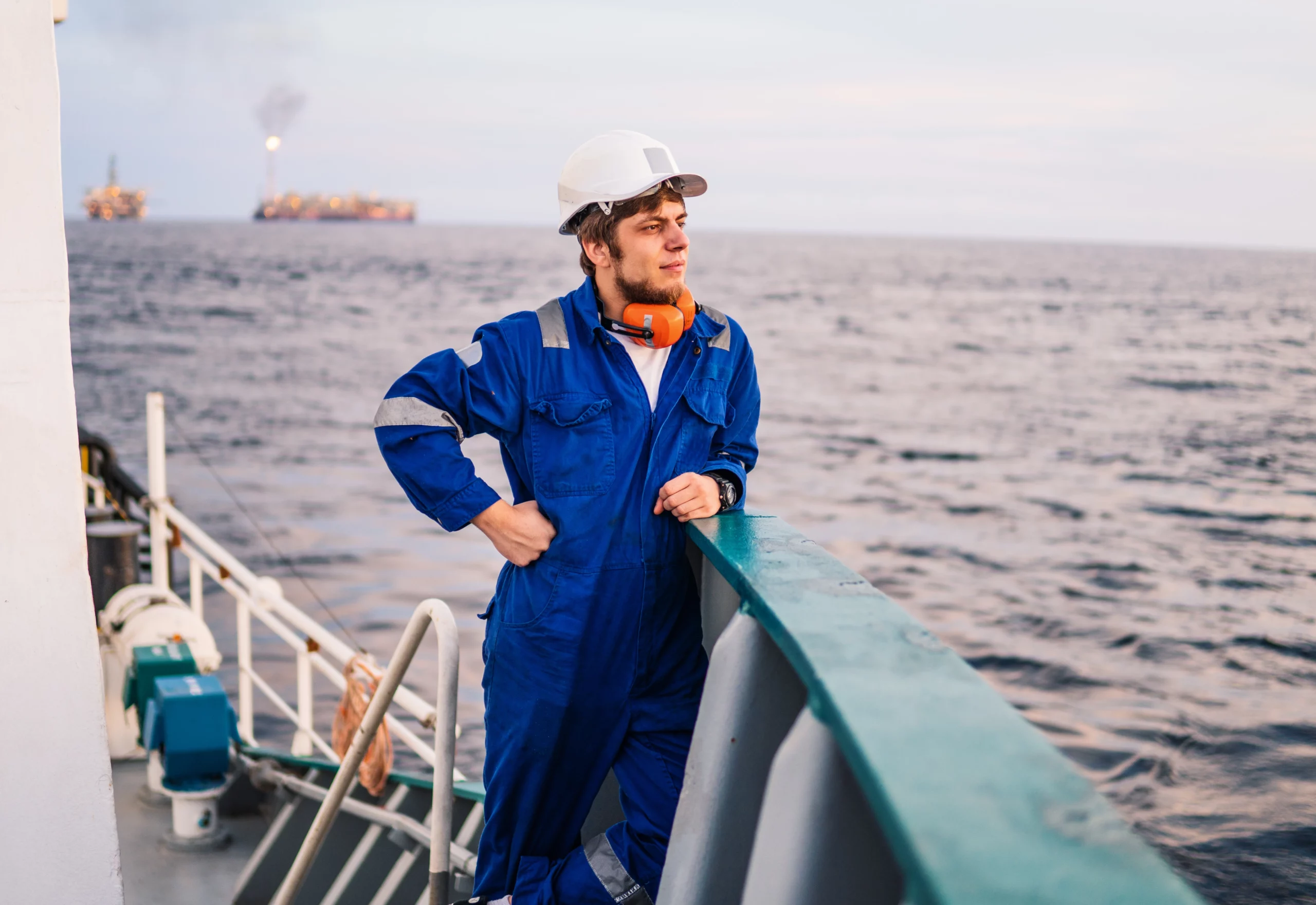 Man in blue coveralls and helmet stands on a ship deck, gazing at the sea with an oil platform in the distance.