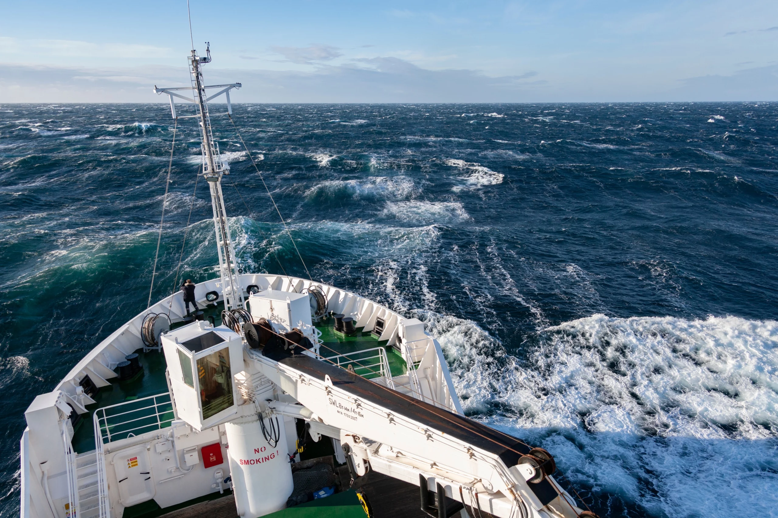 A ship’s bow cutting through choppy blue waves under a clear sky, with a lone figure standing on deck, symbolizing maritime work and challenges.