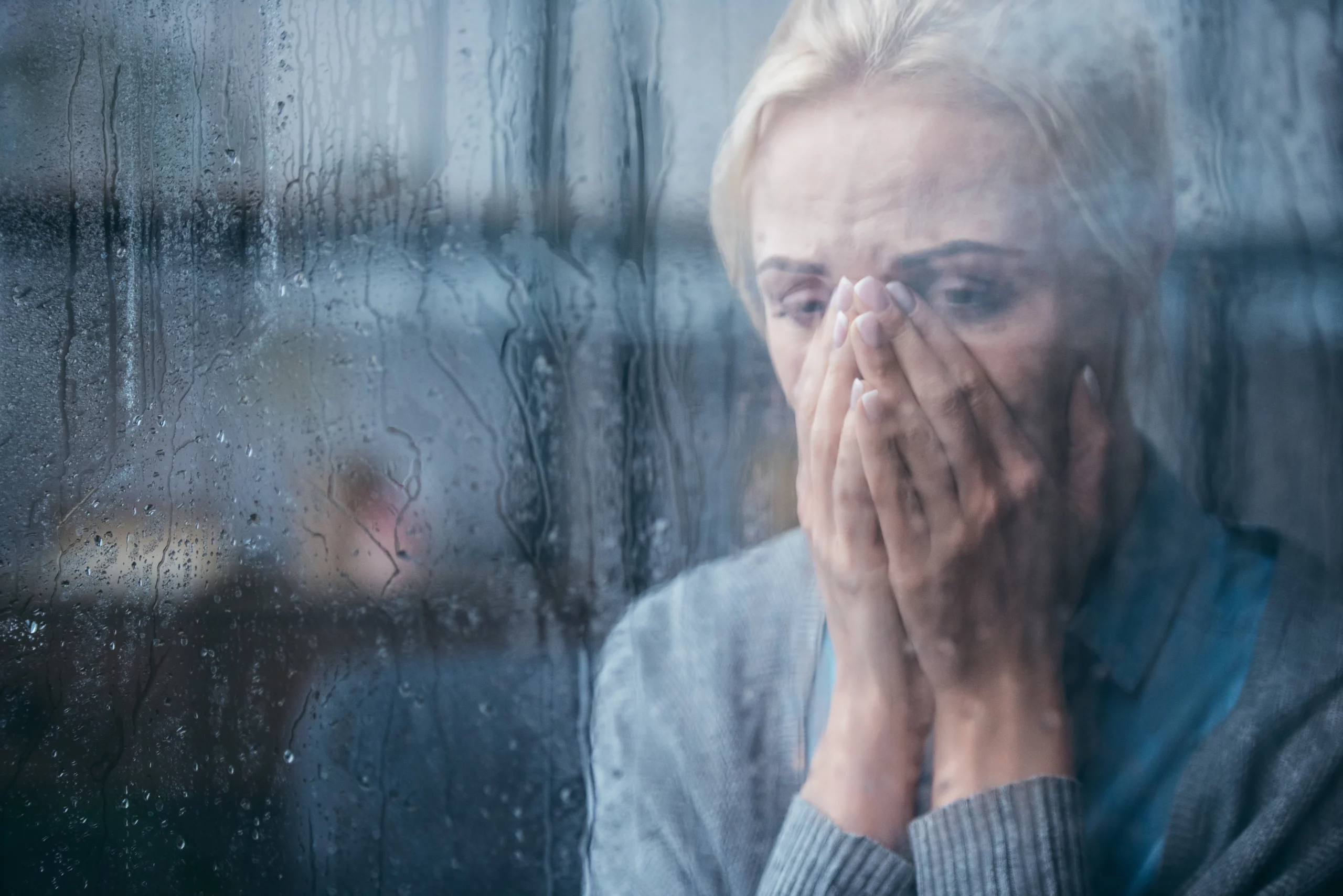 Sad woman behind rain-covered window, hands over mouth
